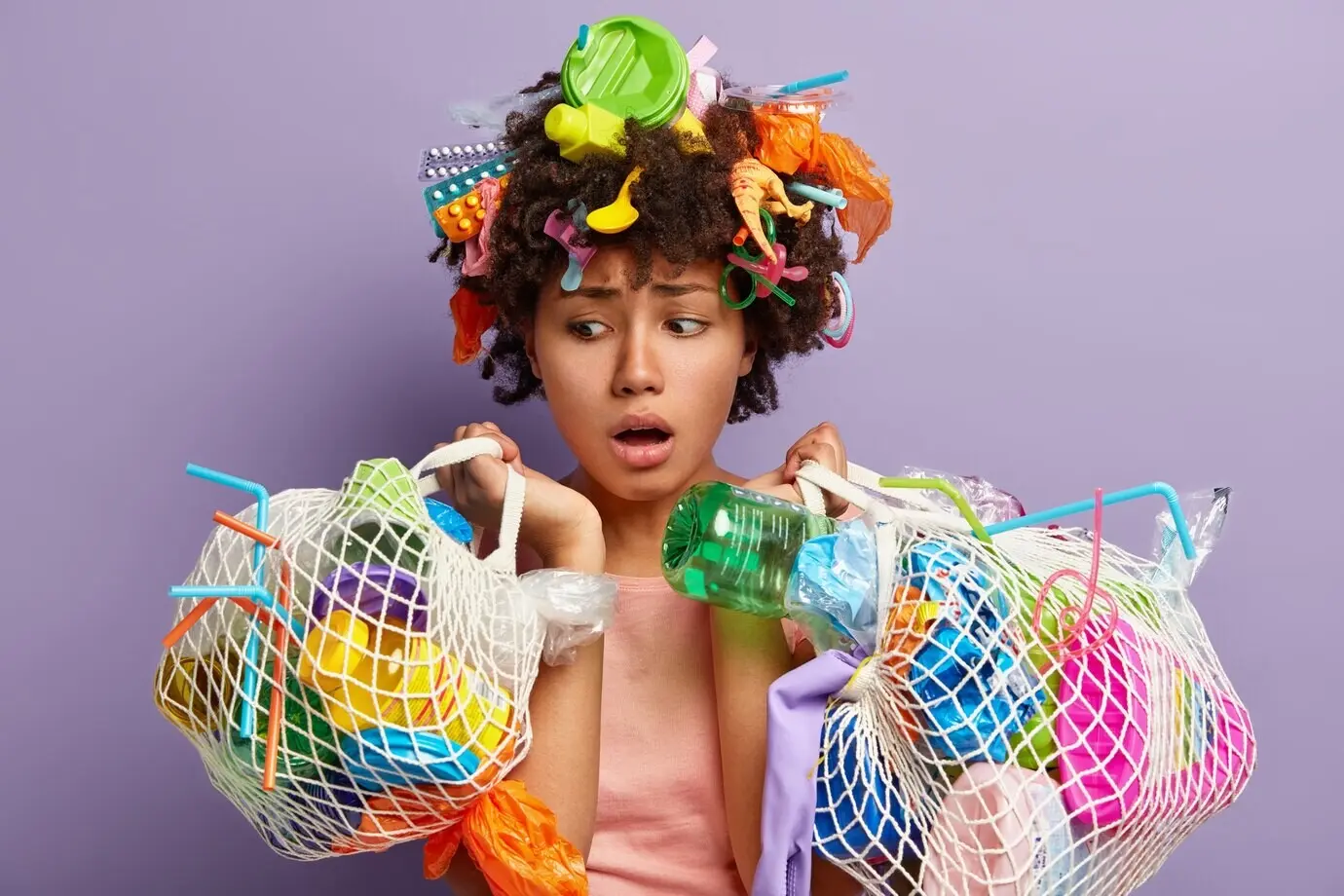 Schockierte dunkelhäutige Frau blickt mit offenen Augen und besorgtem Ausdruck, trägt zwei Netzbeutel mit verschiedenen Plastikabfällen, ehrenamtlich engagiert, isoliert vor einer lilafarbenen Wand.